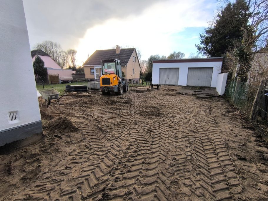 Bauplatz mit Bagger und frisch aufgeschüttetem Sand, im Hintergrund Wohngebäude.