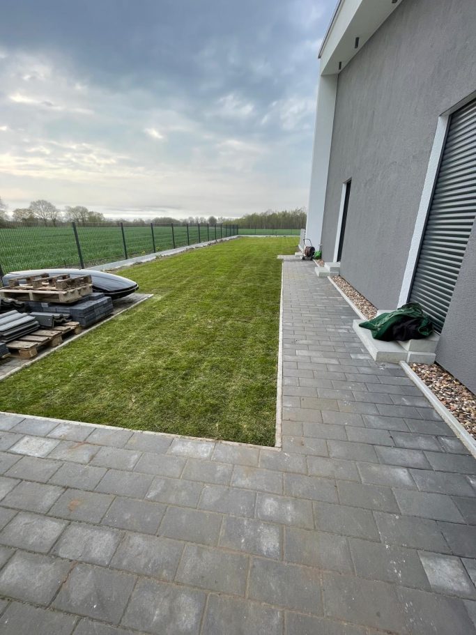 Grüner Rasen entlang einer gepflasterten Terrasse, mit Wolken am Himmel.