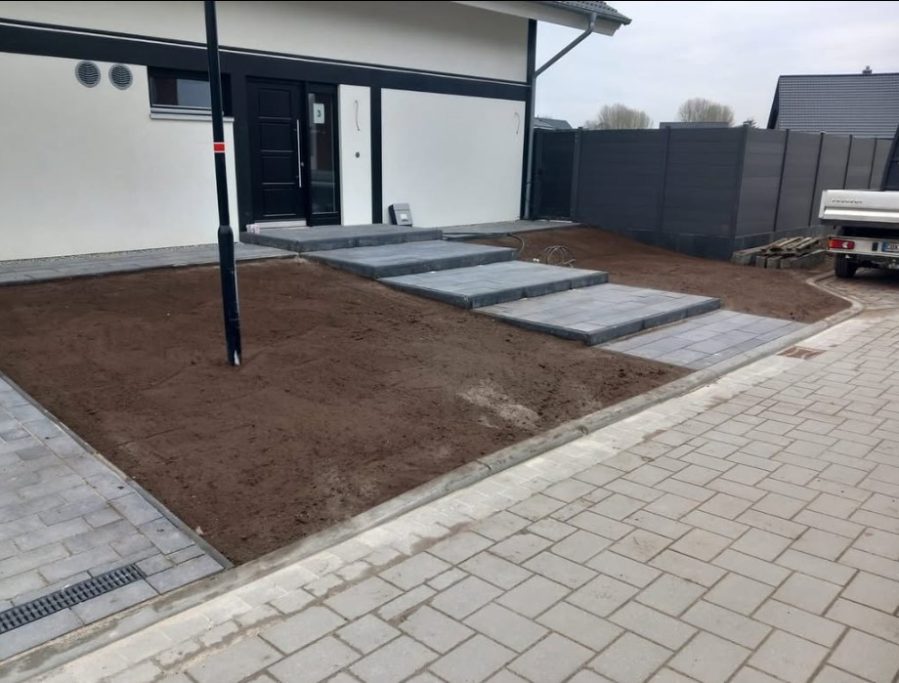 Gartenbereich mit gepflasterten Stufen und unbefestigtem Boden vor einem modernen Haus.